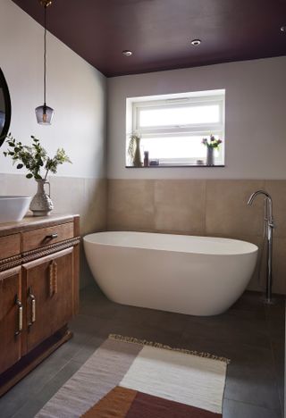 Bathroom makeover: bathroom with large format beige wall tiles to halfway up the wall, large format grey floor tiles, white upper half of the walls and a purple ceiling with spotlights. A white freestanding bath sits under a window