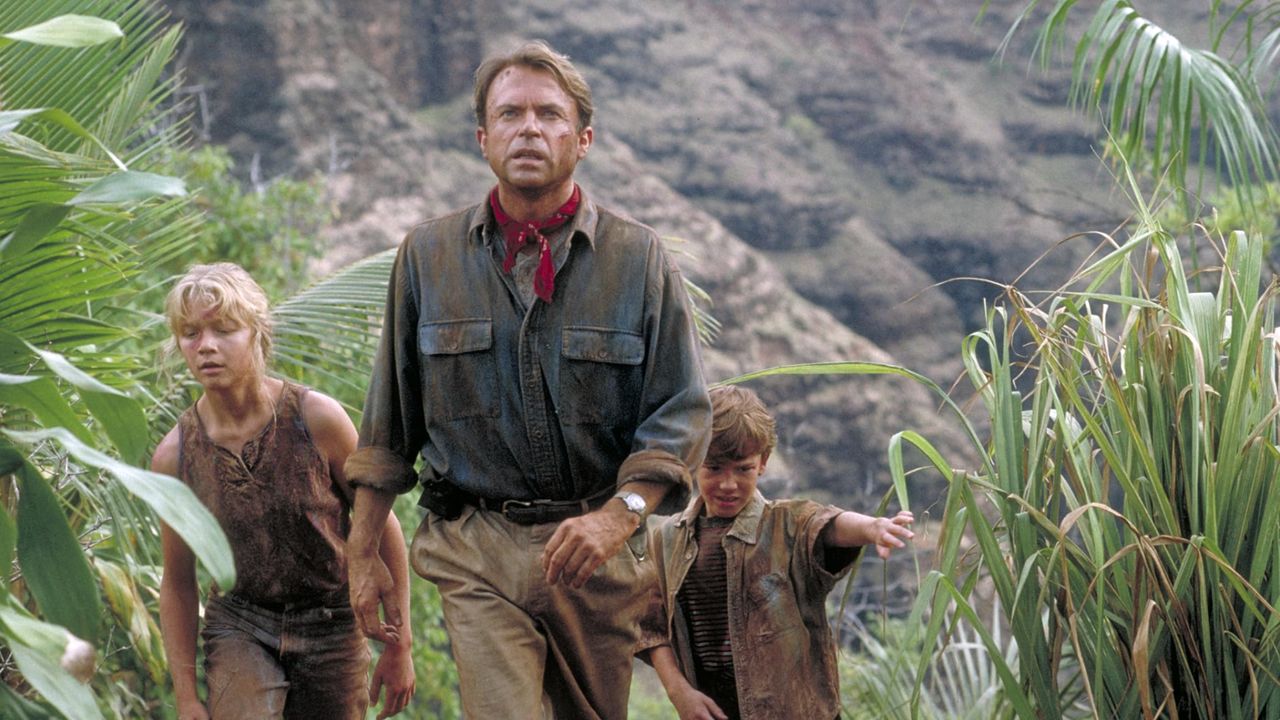 A man wearing a muddy blue shirt and beige trousers with a red neckerchief leads the way in a jungle-type setting for two small children, a girl and a boy, both in muddy clothes