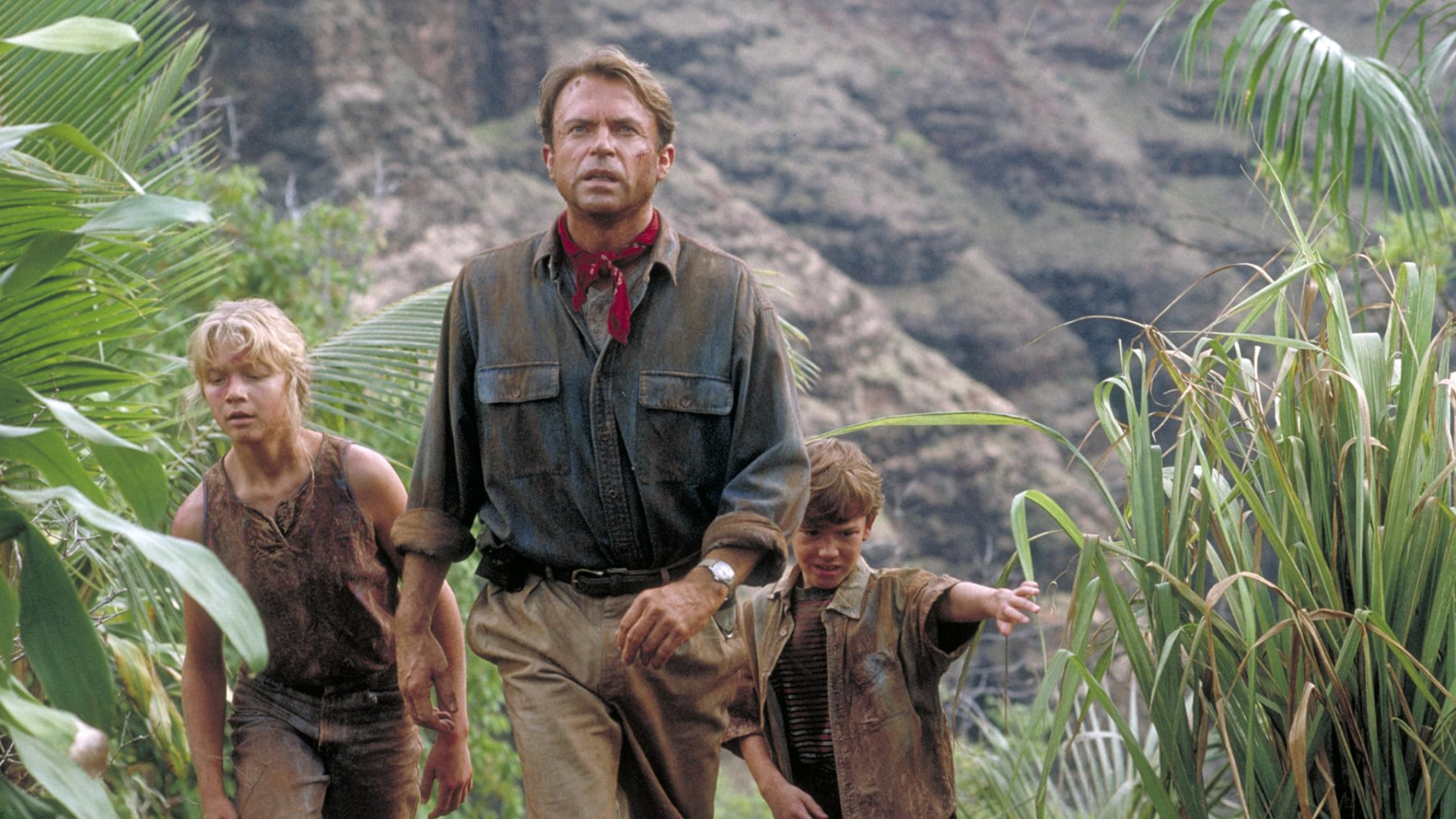 A man wearing a muddy blue shirt and beige trousers with a red neckerchief leads the way in a jungle-type setting for two small children, a girl and a boy, both in muddy clothes