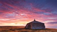 Stone chapel with a cross on top at sunset, surrounded by golden grass under a vibrant pink and purple sky