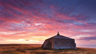 Stone chapel with a cross on top at sunset, surrounded by golden grass under a vibrant pink and purple sky