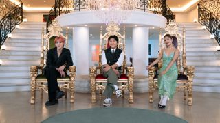 three people sitting in golden thrones sit in front of an opulent staircase