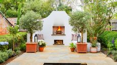 Secret hideaway Mediterranean style garden with white stone walls and seating, open fire place, log storage, grey cushions, flag stone patio with pond, lanterns, hydrangea and trees in terracotta planters