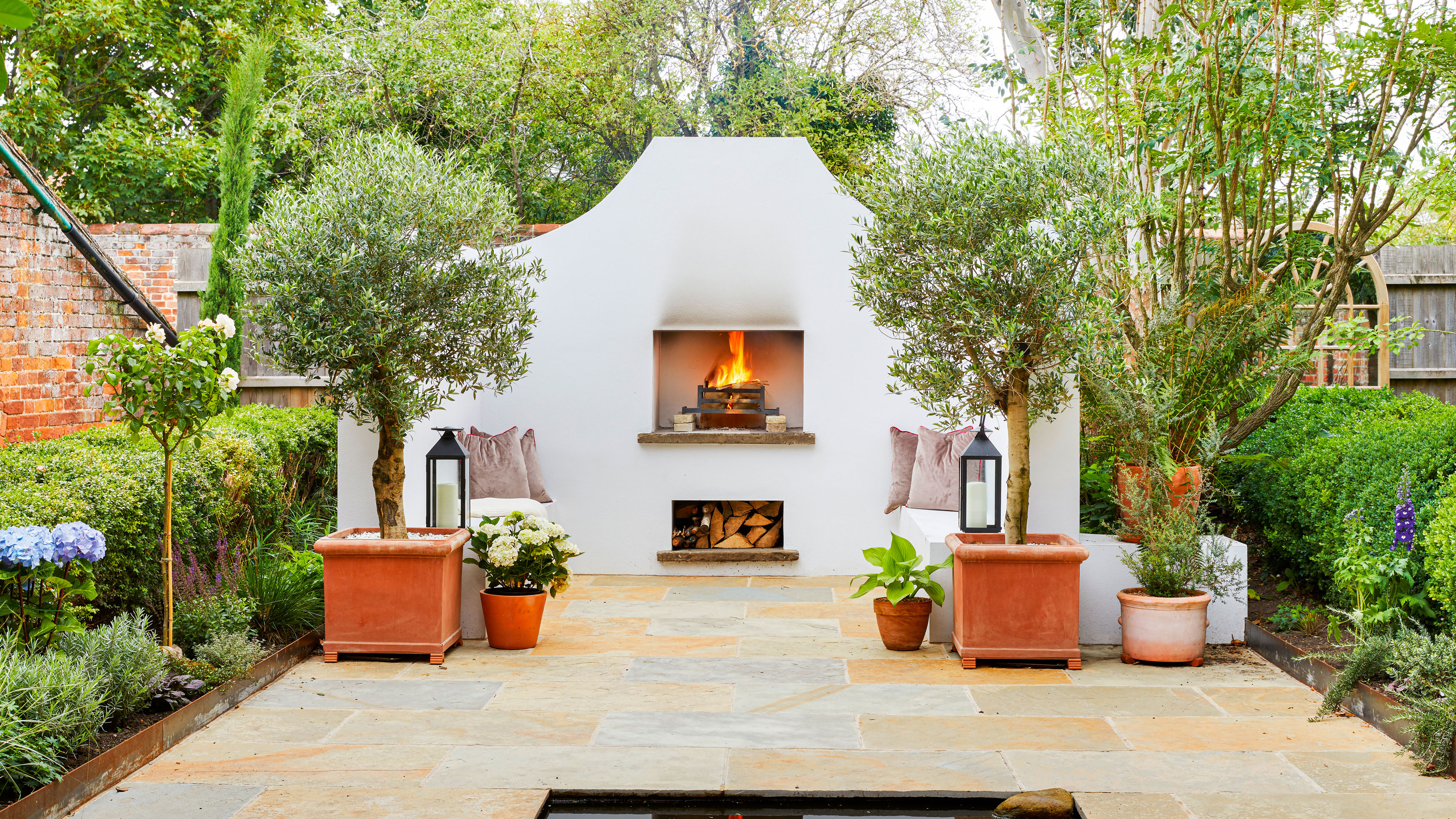 Secret hideaway Mediterranean style garden with white stone walls and seating, open fire place, log storage, grey cushions, flag stone patio with pond, lanterns, hydrangea and trees in terracotta planters