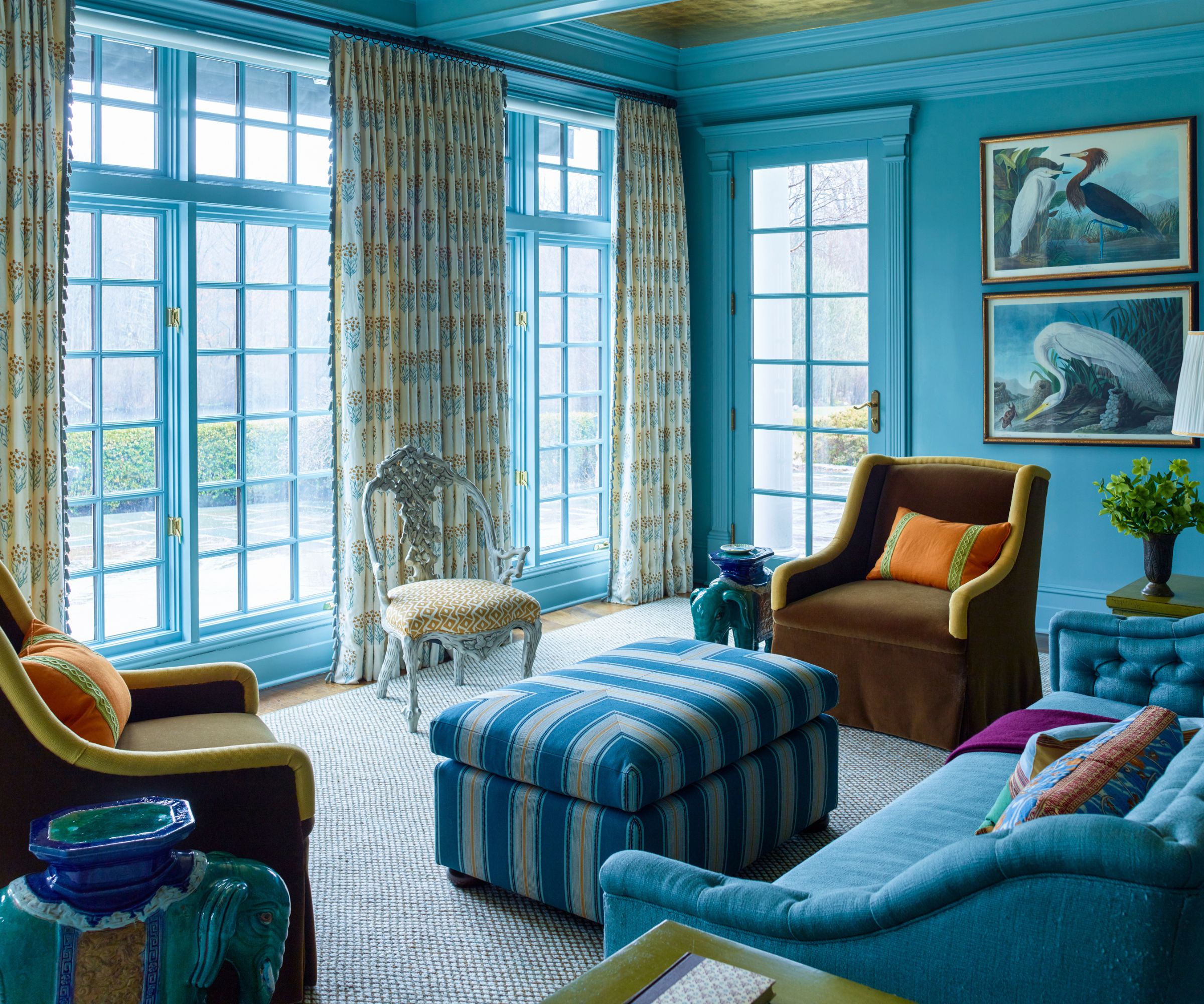 Living room with bright blue walls, large French doors, a blue sofa, a blue striped ottoman, two brown velvet armchairs opposite one another, an elephant side table and two prints of herons