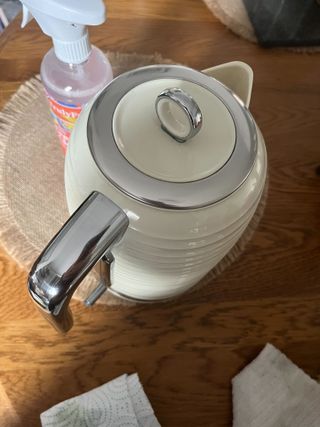 A cream kettle with stainless steel trims on a wooden table next to a cleaning bottle and some cloths