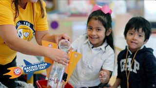 Two children next to an adult smile as liquid is poured in an bottle.