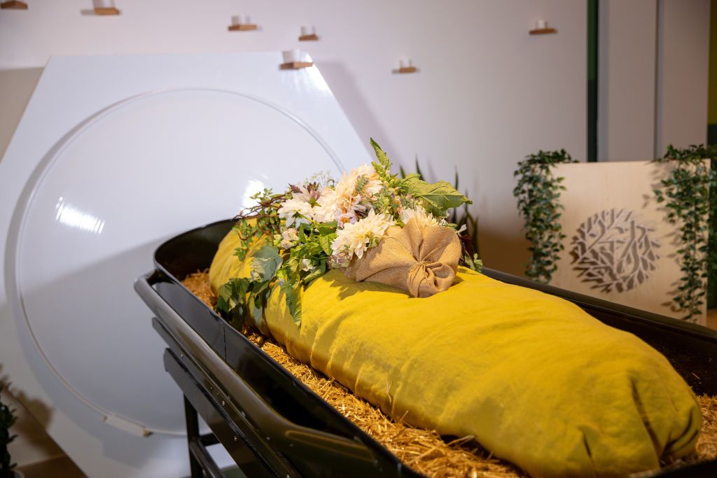 A shrouded mannequin covered with flowers rests near a composting vessel.