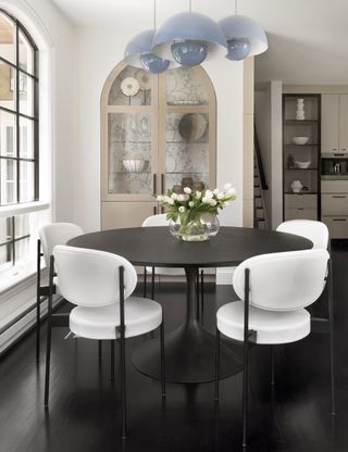 A dining area with a white circular table and large windows with black trim and a display cabinet in the background