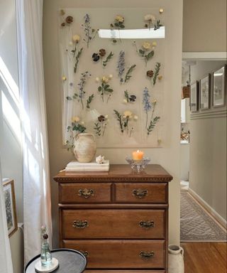 An entryway with a wall art frame with plant cuttings in it and a dark brown wooden drsser with candles on top of it