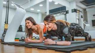 Two women taking part in EMS training doing a forearm plank on the floor of a gym