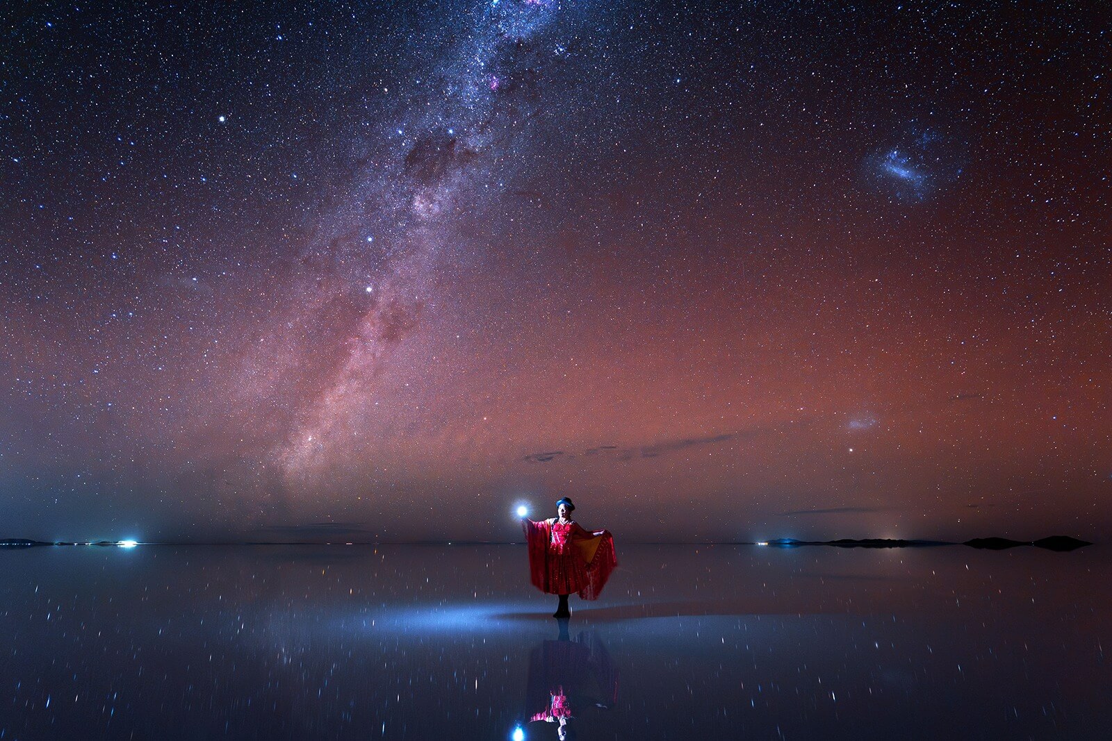 Woman holds lamp while standing on watery surface reflecting starry night sky.