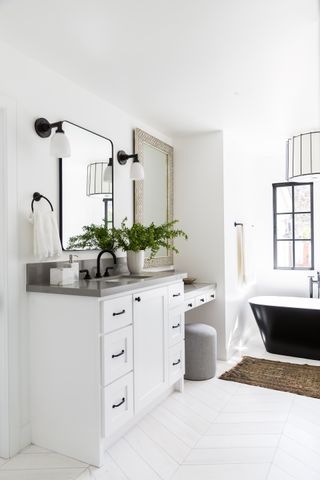 white bathroom with split level vanity with sink one side, make up vanity the other. two mirrors, white floor, rug, black bath, black crittall windows