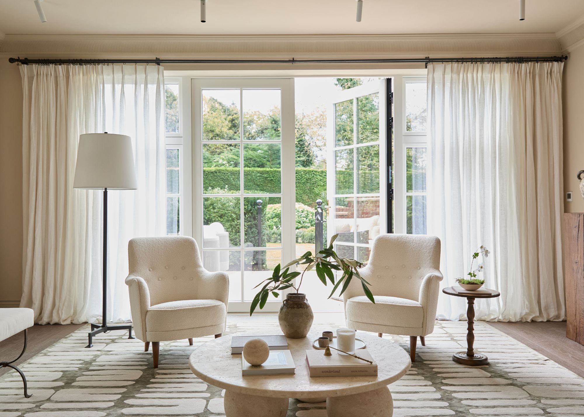 A neutral living room with modern decor, large white French doors leading to the garden, and white sheer curtains.