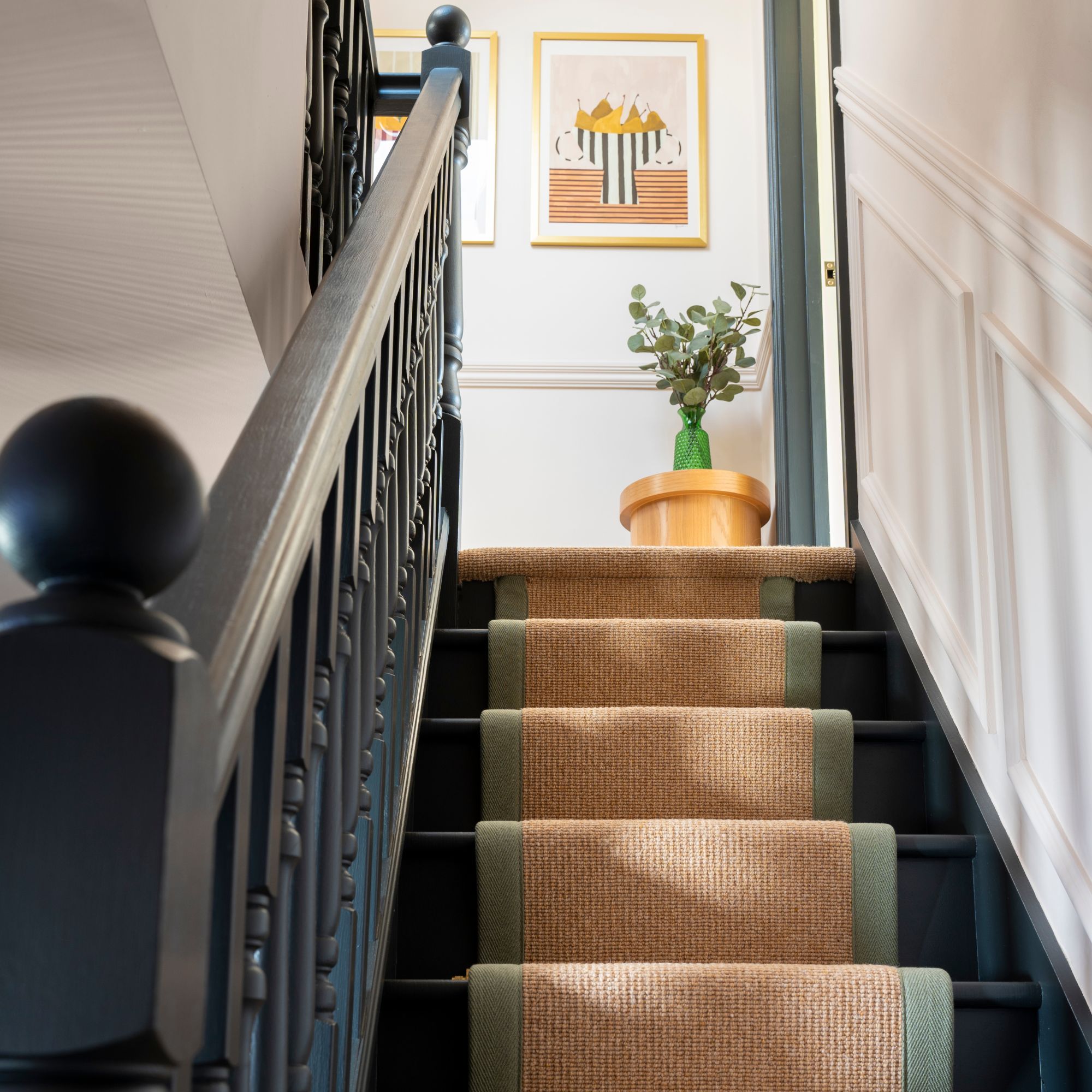 Black wooden staircase with a wool carpet runner, and a vase of flowers and prints on the wall at the top of the landing