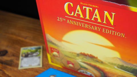 Catan 25th Anniversary Edition box on a wooden table, with cards and the board beside it