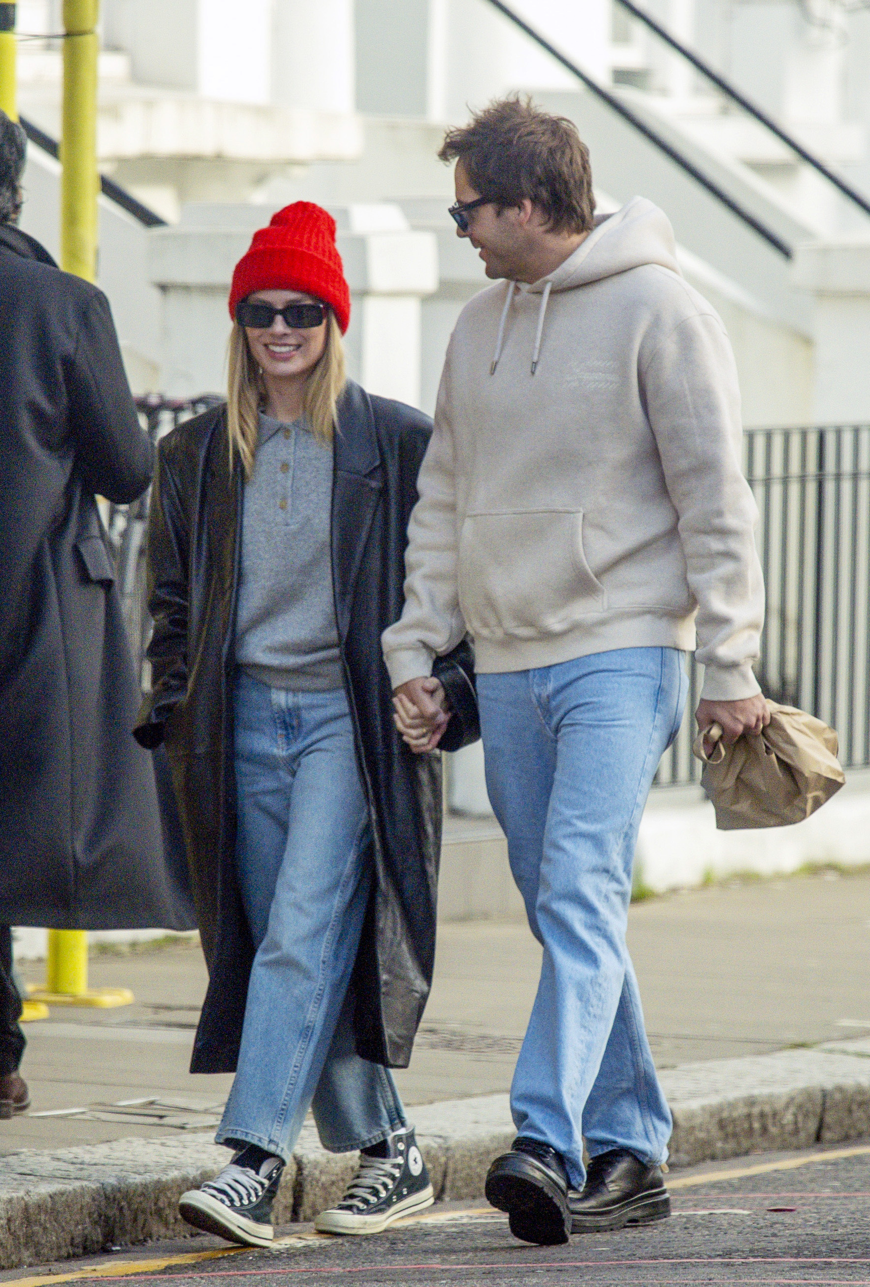 Margot Robbie walks down the street in Notting Hill, London, wearing cropped jeans with Converse and a grey knit with a long-line leather jacket and red beanie.