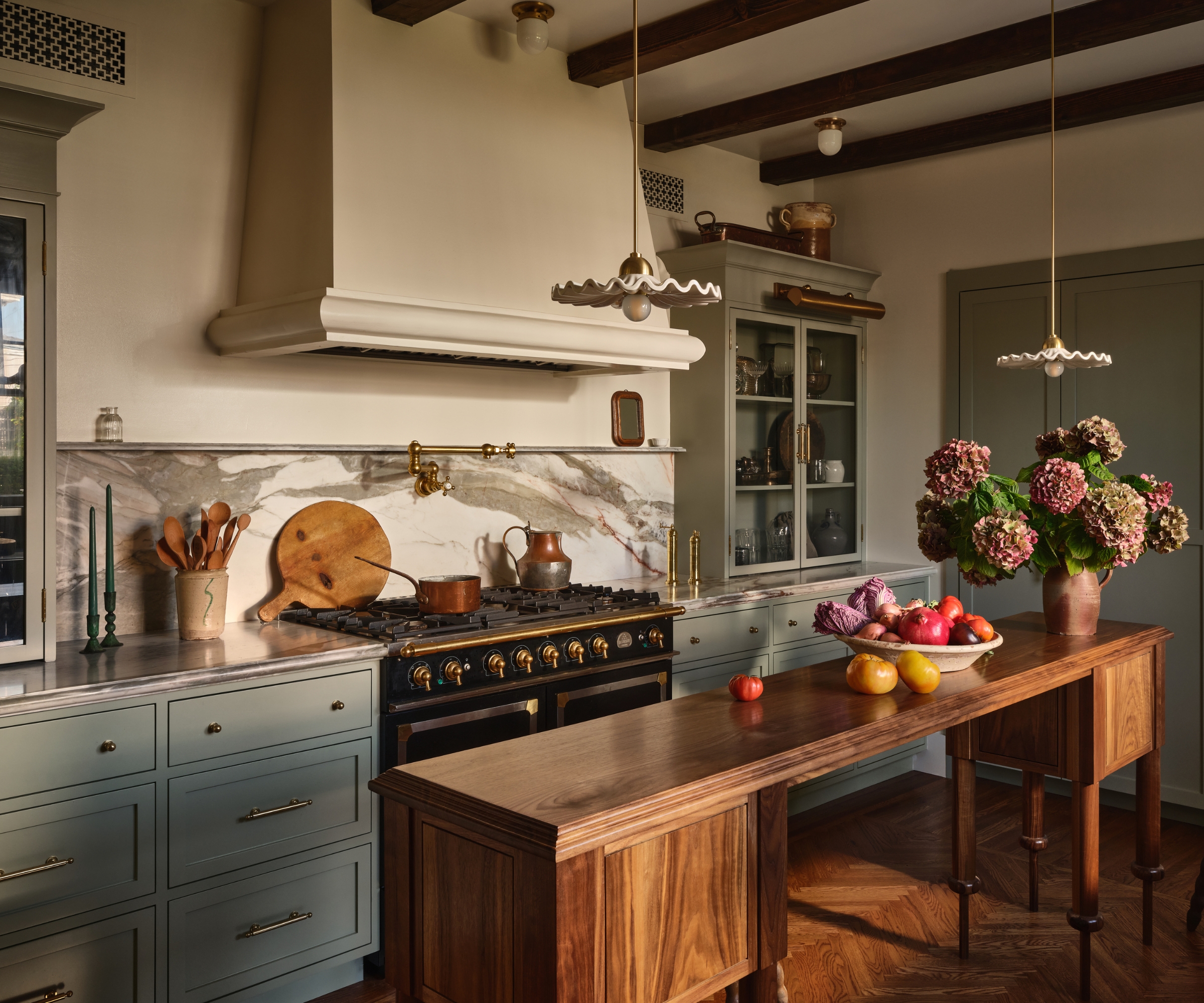 A small kitchen with light blue cabinets, warm white walls, a custom plaster range hood, a black range cooker, marble countersplash, a custom narrow wooden island, and collected decor