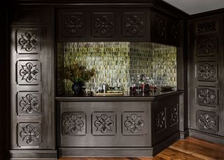 A dark wooden home bar carved with a gothic floral motif with a green zellige tiled wall
