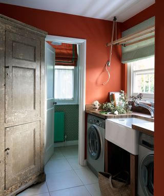 A bright orange laundry room with a white basin and brown wooden countertops with matching washer and dryers and an antique wooden cabinet, and open door to a green wallpapered room behind with a turquoise radiator and orange and green open blind