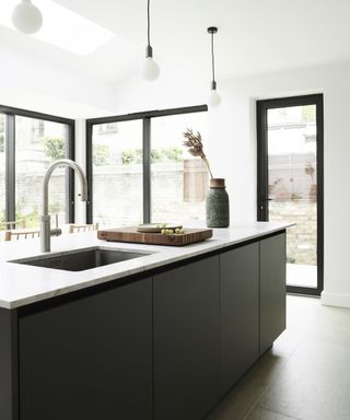 A modern kitchen with white walls, black framed floor-to-ceiling windows and grey island with quartzite worktop and sink