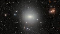 A black background of space is speckled with a wide array of stars. At the center is a flat, glowing galaxy shaped like a stretched out sombrero. 