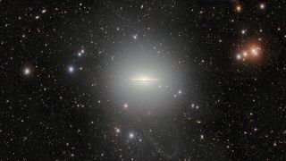 A black background of space is speckled with a wide array of stars. At the center is a flat, glowing galaxy shaped like a stretched out sombrero. 