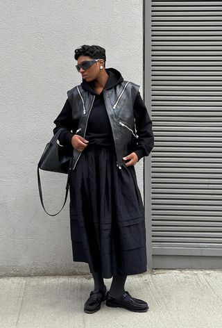 A woman standing outside wearing a black hoodie under a leather vest with a black skirt and black loafers.