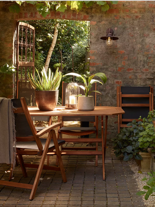 a rustic outdoor paved garden with a wooden table, plants and lights, and a brick wall with an iron wrought door