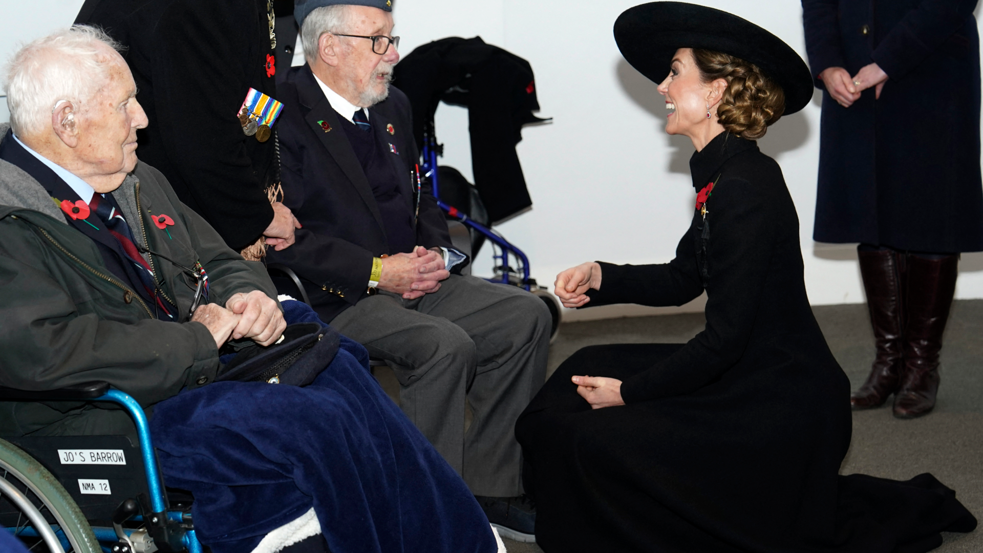 Kate Middleton kneeling in a black coat and hat talking to veterans in wheelchairs