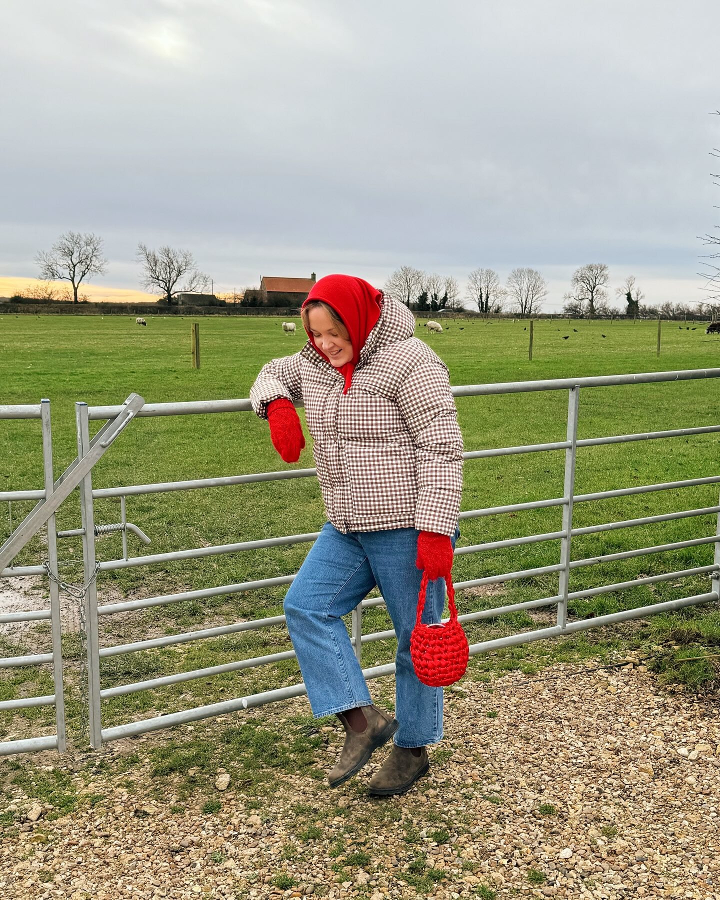 Woman wears a brown gingham puffer jacket, brown suede ankle boots, straight leg jeans and a red head scarf, mittens and bag.