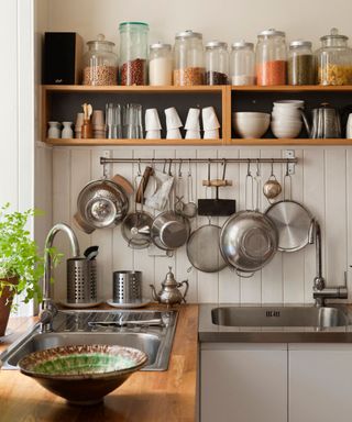 A kitchen with two wooden shelves with dried goods jars, cups, and glasses on top, a paneled wall with silver cookware hanging from it, and a wooden counter with a silver sink