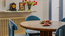 modern dining room with round table, blue dining chairs, a pendant light overhead, yellow radiator, and floral floating shelf with clock, candles, picture, and vase of flowers