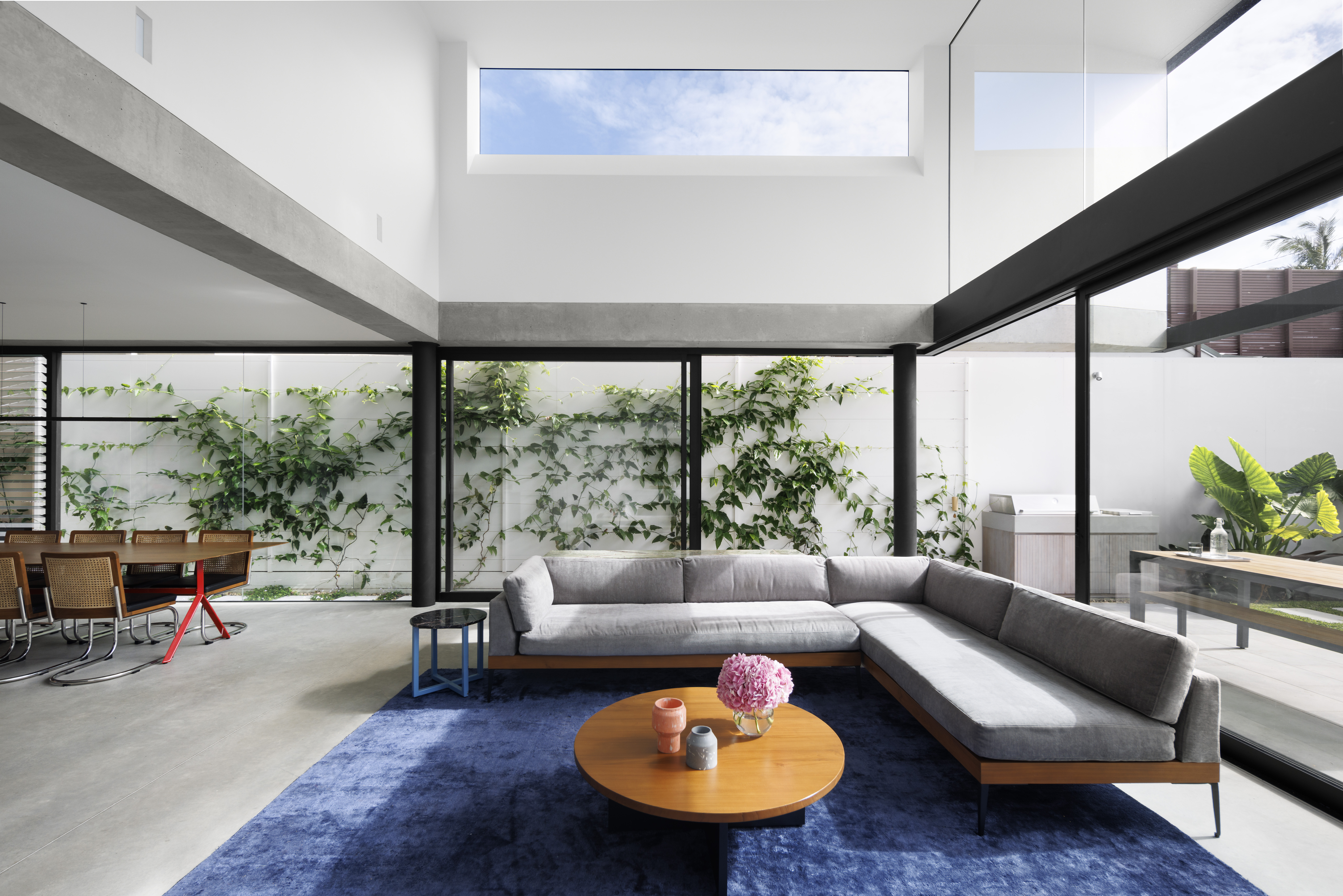 A bright modern open-plan living room with high ceilings, grey sectional sofa on a blue rug, round wooden coffee table, and large glass sliding doors looking out to a ivy-covered wall.