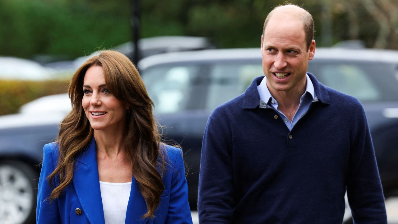 Kate Middleton wearing a blue blazer and Prince William wearing a blue sweater walking through a parking lot 