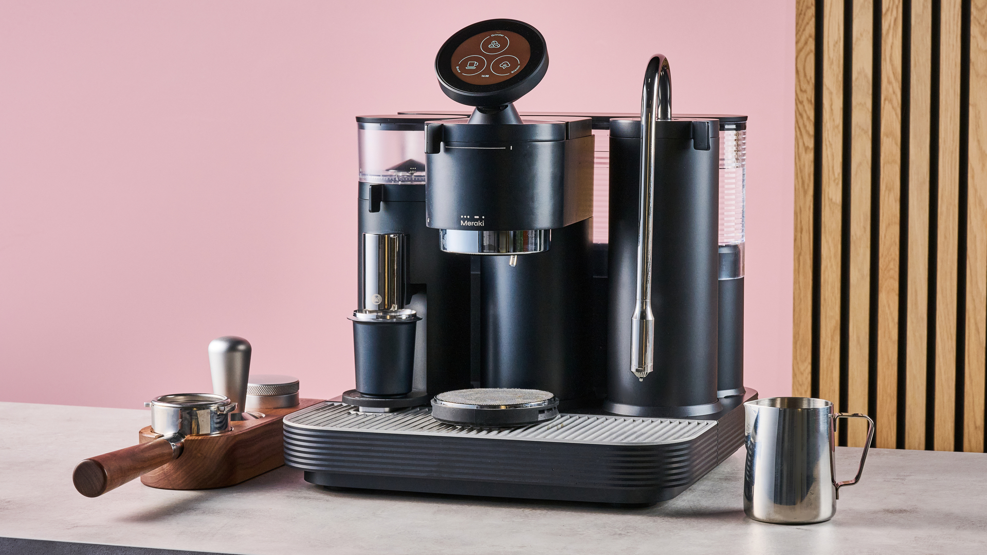 The Meraki Espresso Machine with a tamp, coffee distributor and portafilter to its left and a milk jug to its right, in front of a pink and wooden effect background.