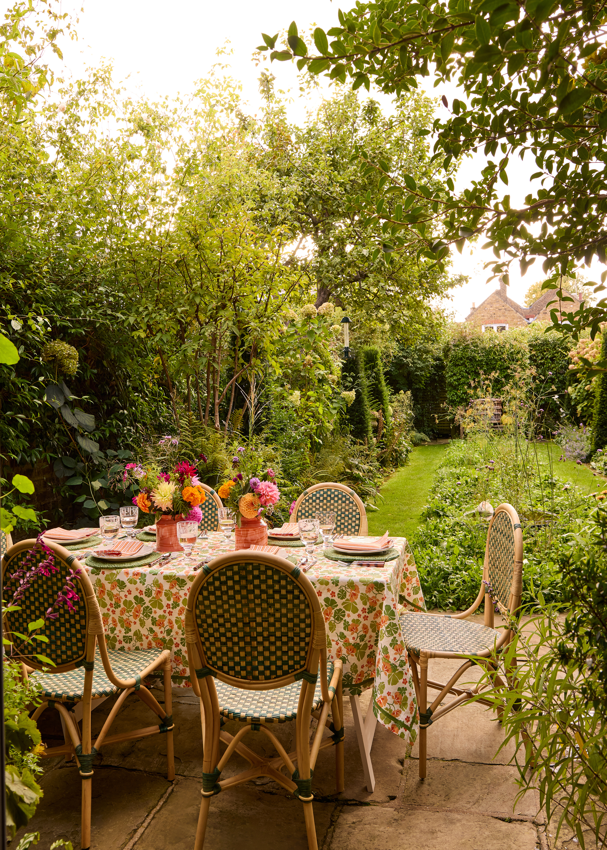 An outdoor dining table with a patterned tablecloth, two vases of flowers, and dinnerware sets