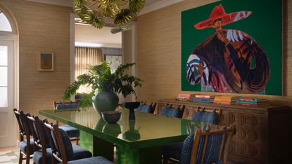 Dining room with glossy green table, blue upholstered chairs, jute walls, vintage wood sideboard and colorful artwork.