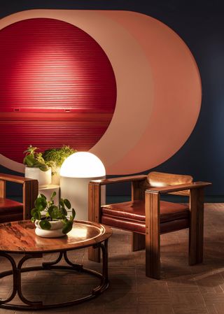 A suffused atmosphere brought to life by a semi-spherical standing light illuminates the wood and leather surfaces of a hotel lobby, decorated with geometric colorful shapes.