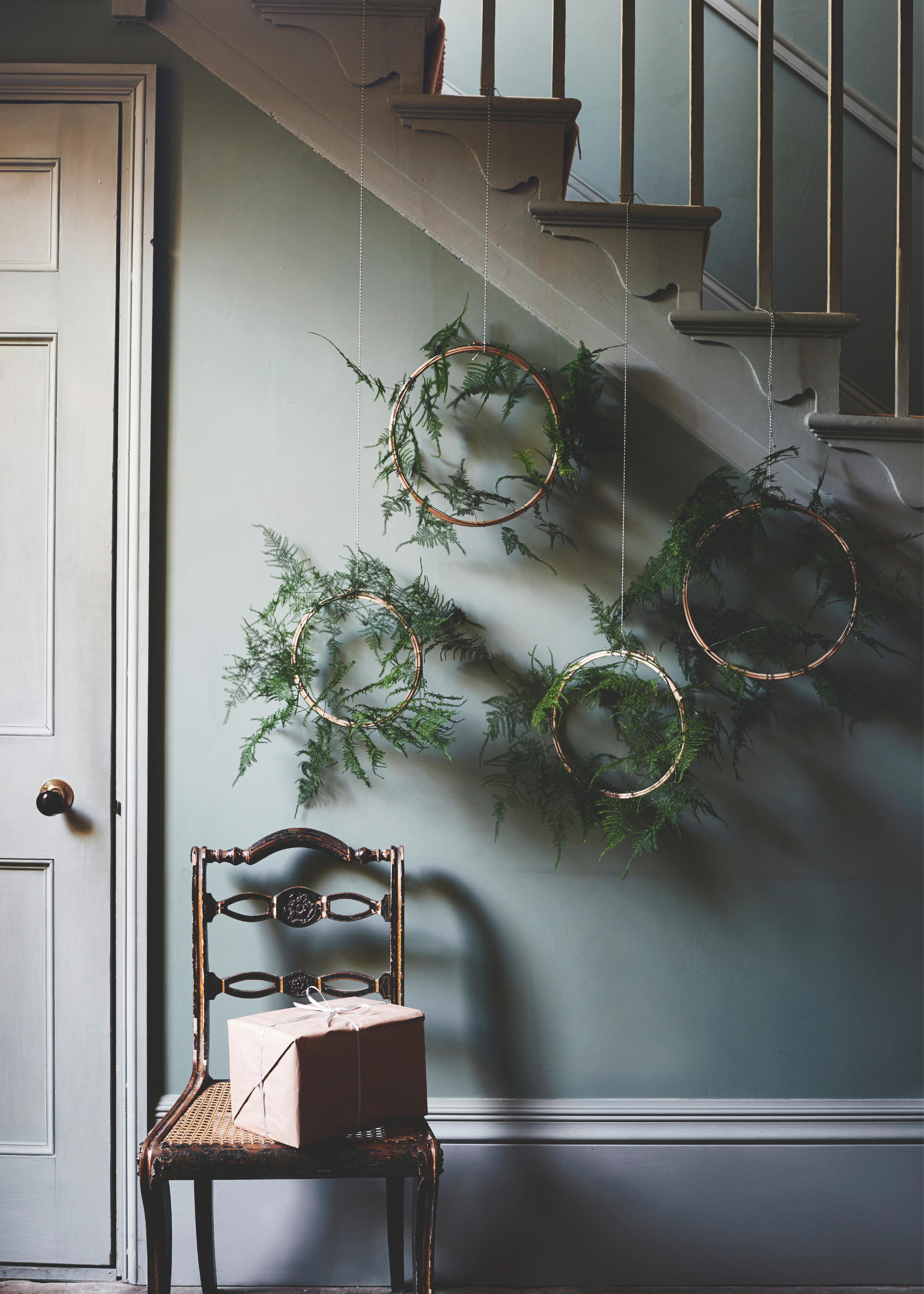 Four wreaths crafted from embroidery hooks covered in ferns are hung at varying heights off the side of the staircase above a chair in a hallway