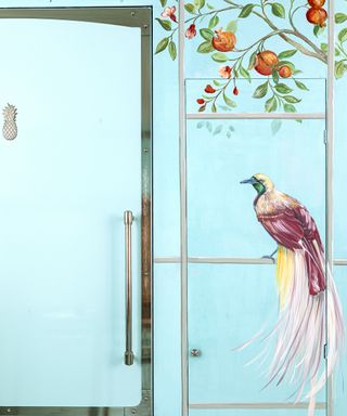 The built-in fridge of a turquoise kitchen with hand painted bird and tree illustration