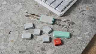 A photo of some spare keycaps and a keycap/switch puller included with the NuPhy Air75 V2 on a stone slab with a blue wall in the background.