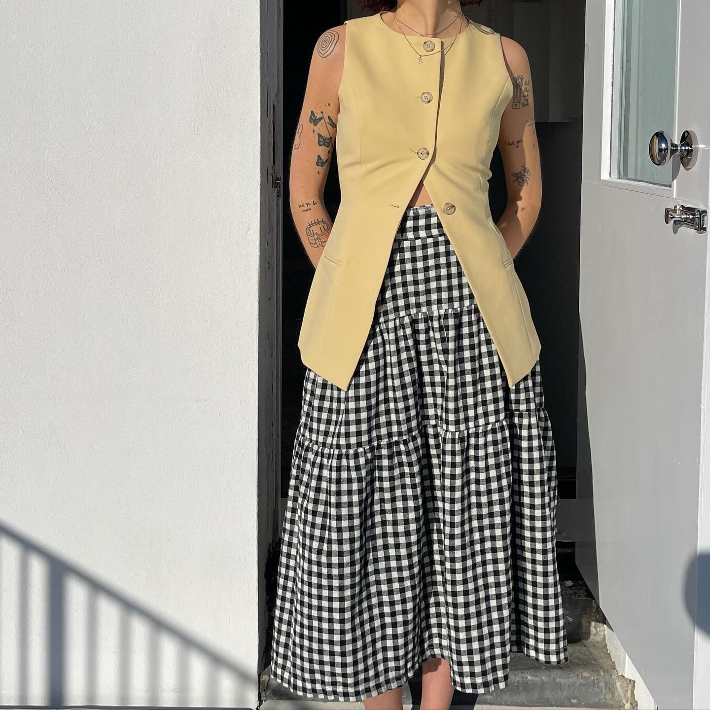 Fashion person Evie wears a butted yellow waistcoat and a black and white drop waist gingham skirt. She is stood in front of a white open door with a white wall on the other side in a recent image from her Instagram.