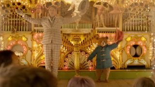 Hugh Grant stands with Paddington Bear on a stage, both holding their hats in the air in Paddington 2.