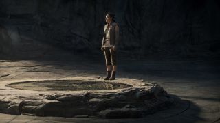 A young woman standing in a cave next to a circular indentation on the floor.