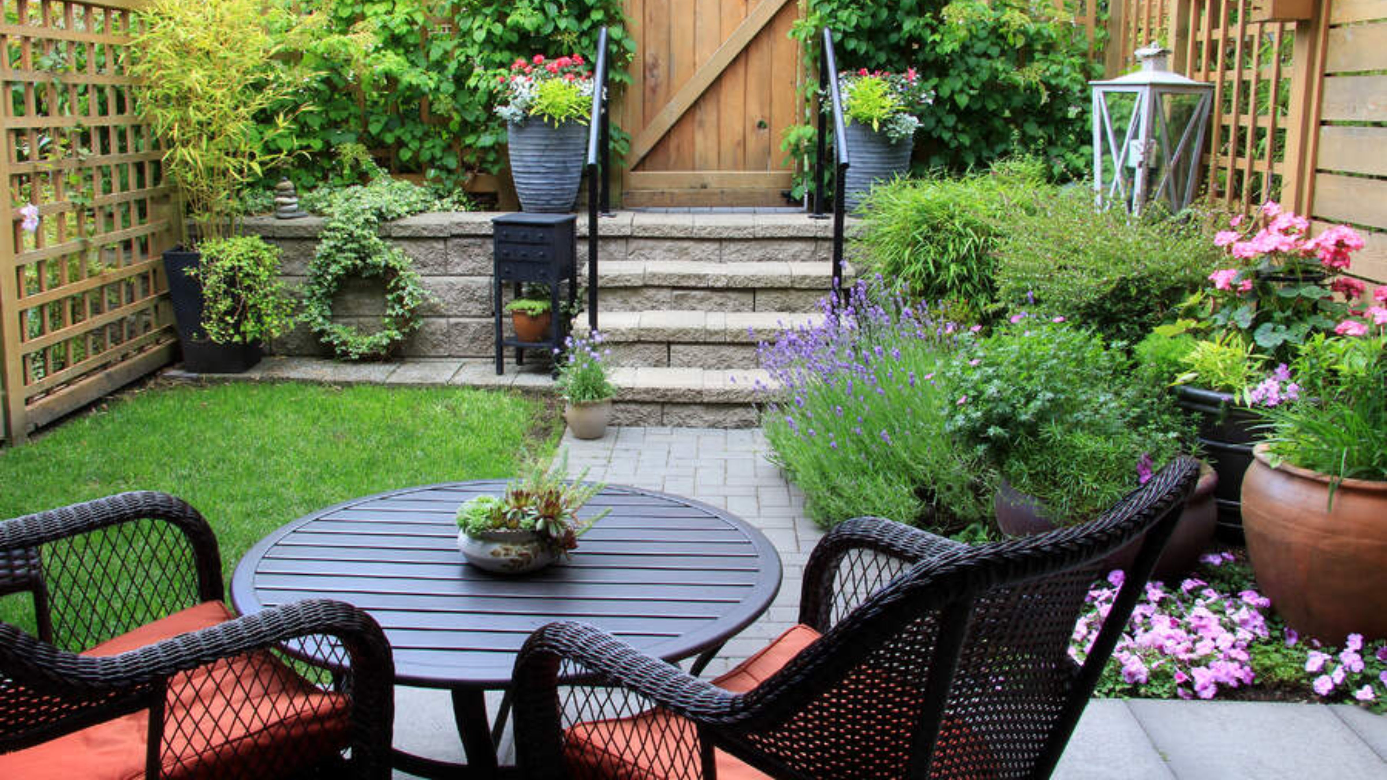 Petite backyard area featuring a small garden, stone steps, and a black woven bistro set with brick red cushions.