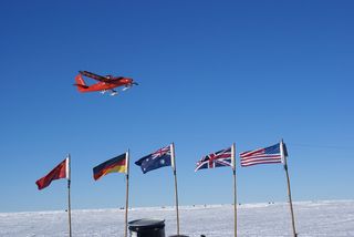 A Twin Otter aircraft flies over flags at an Antarctic outpost.