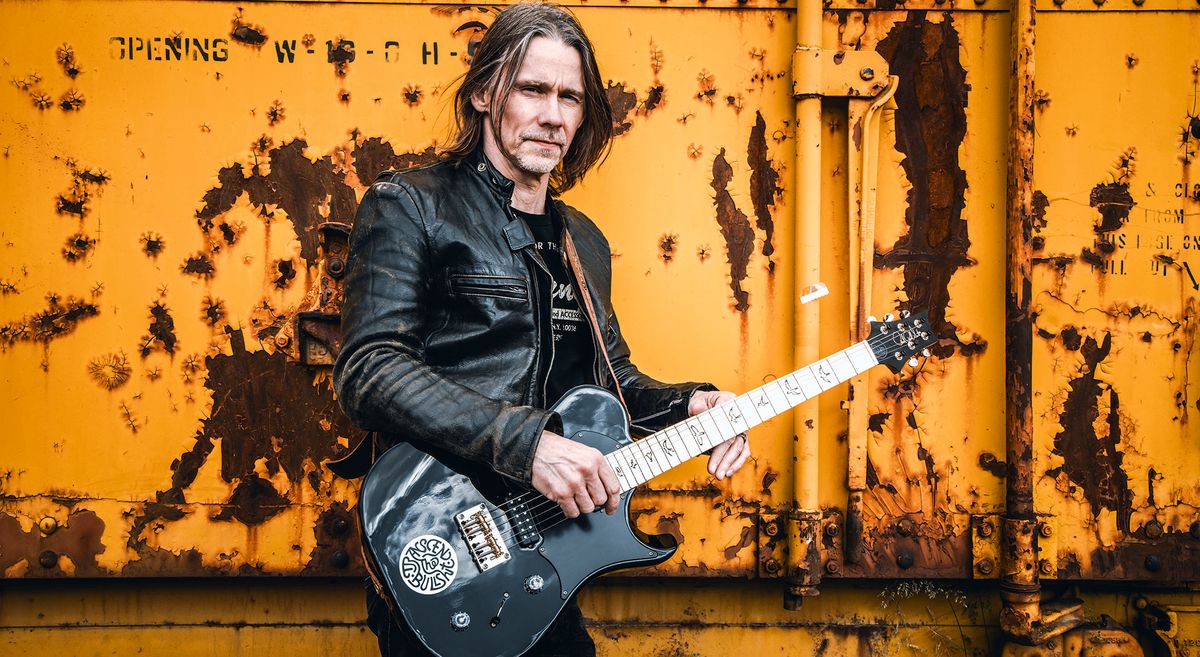 Myles Kennedy stands in front of a rusted orange container. He wears a black leather jacket and has his signature PRS guitar.