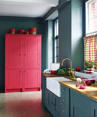 A blue grey kitchen with bright red painted freestanding cabinet and colorful gingham blinds.