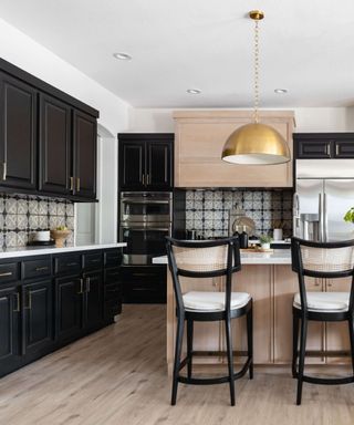 black kitchen with some light wood wall units and island, black and gray backsplash tiles, black bar stools, brass pendant lights, hardwood floor, white countertops